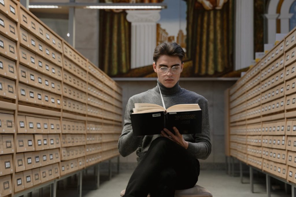 A young man reads a book in a modern library archive, surrounded by card catalogs.